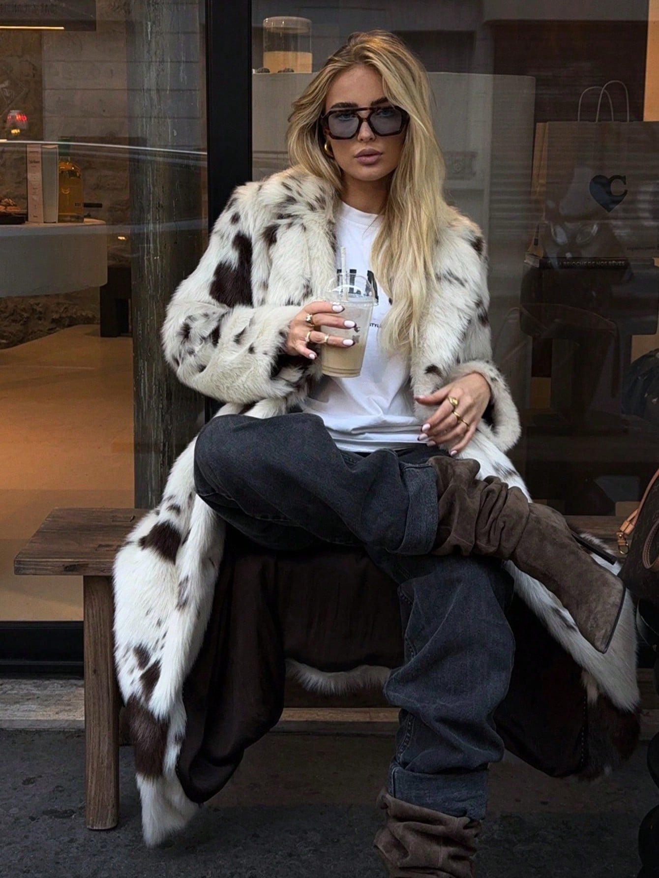 Woman wearing a stylish Long Faux Fur Effect Coat with sunglasses, sipping a drink in a chic setting