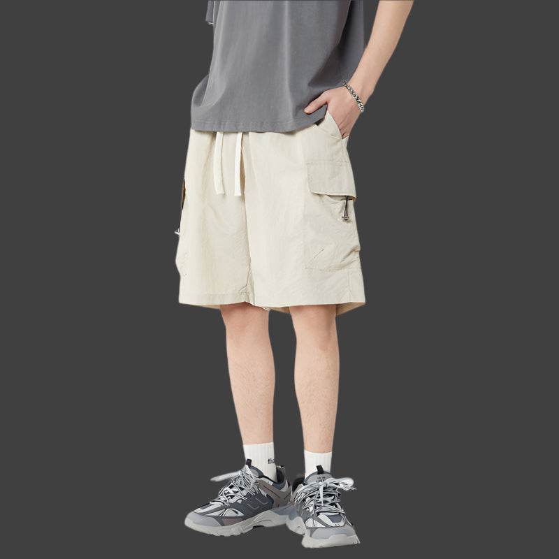 Pantaloni scurți casual cu buzunare multiple bej, purtați de bărbat cu tricou gri și pantofi sport gri
