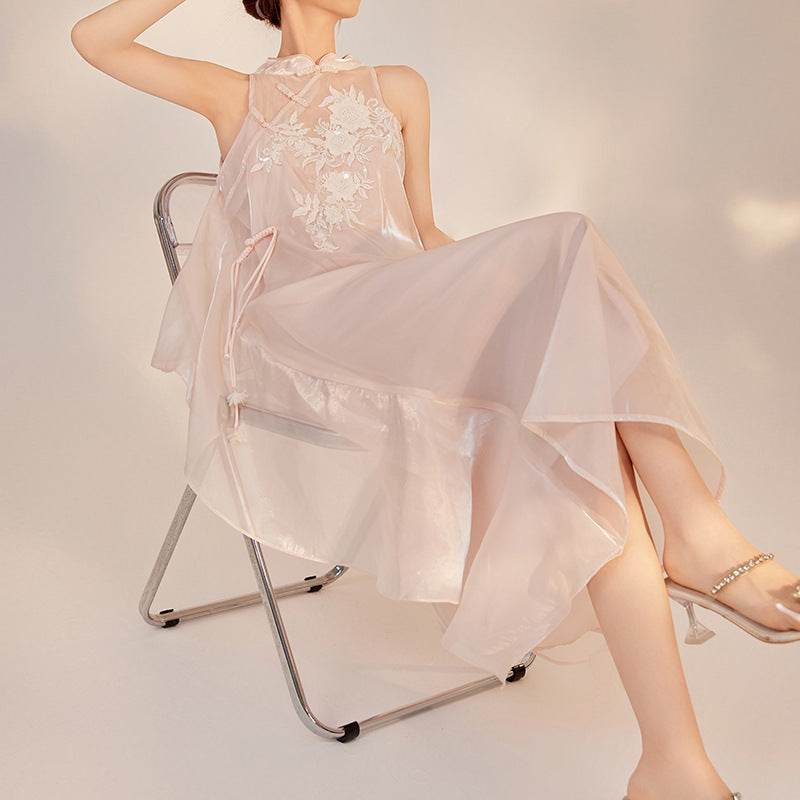 Femeie purtând rochie lungă cu broderie albă elegantă, așezată pe scaun metalic pliabil